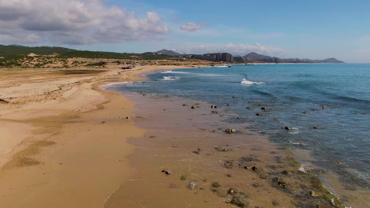At 0:03, the aerial angle shows a wider stretch of El Tule Los Cabos beach, exposed sand in the foreground, rocks in the shallows, surf bands offshore, and no visible people.