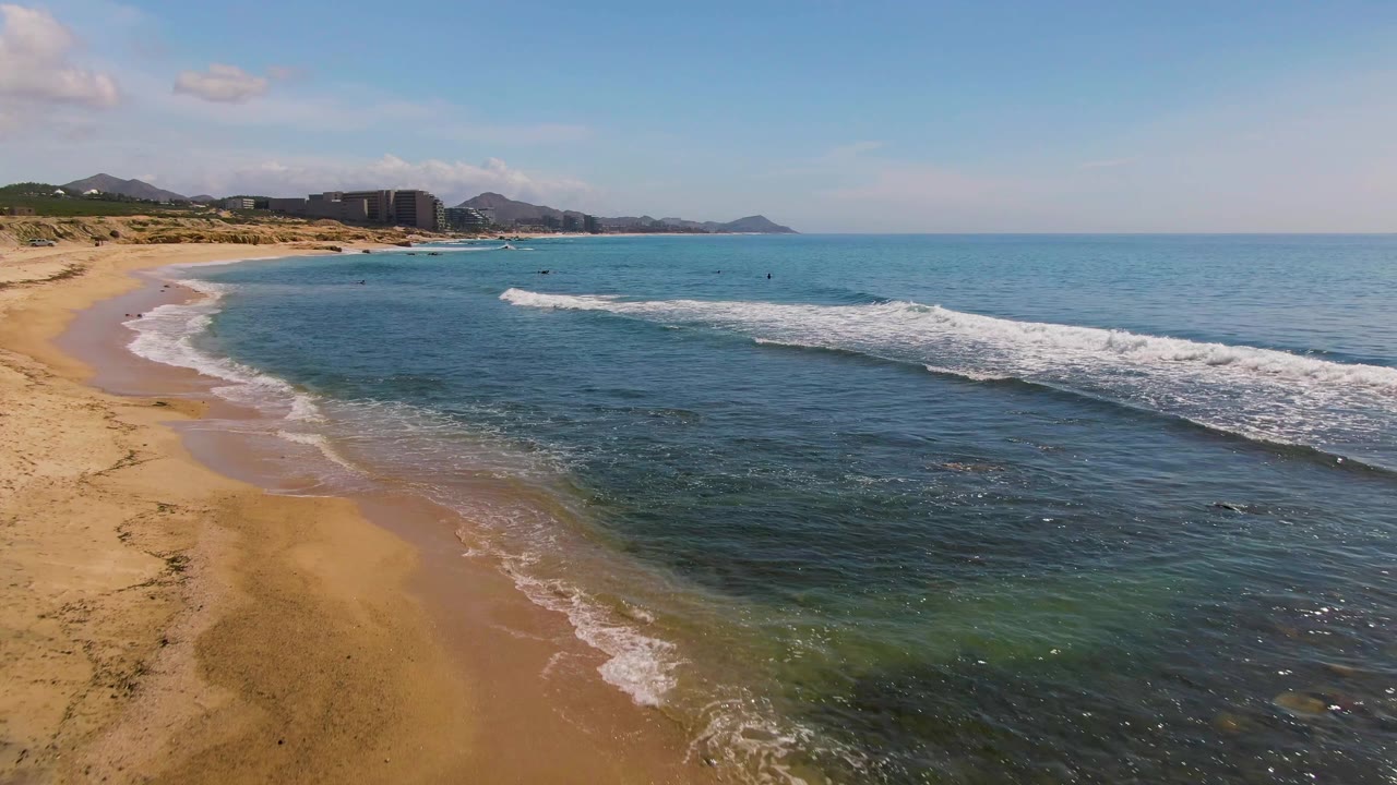 Aerial view at El Tule Los Cabos showing sandy beach on the left, clear blue-green water, a low white wave line, distant shoreline development, and no visible people.