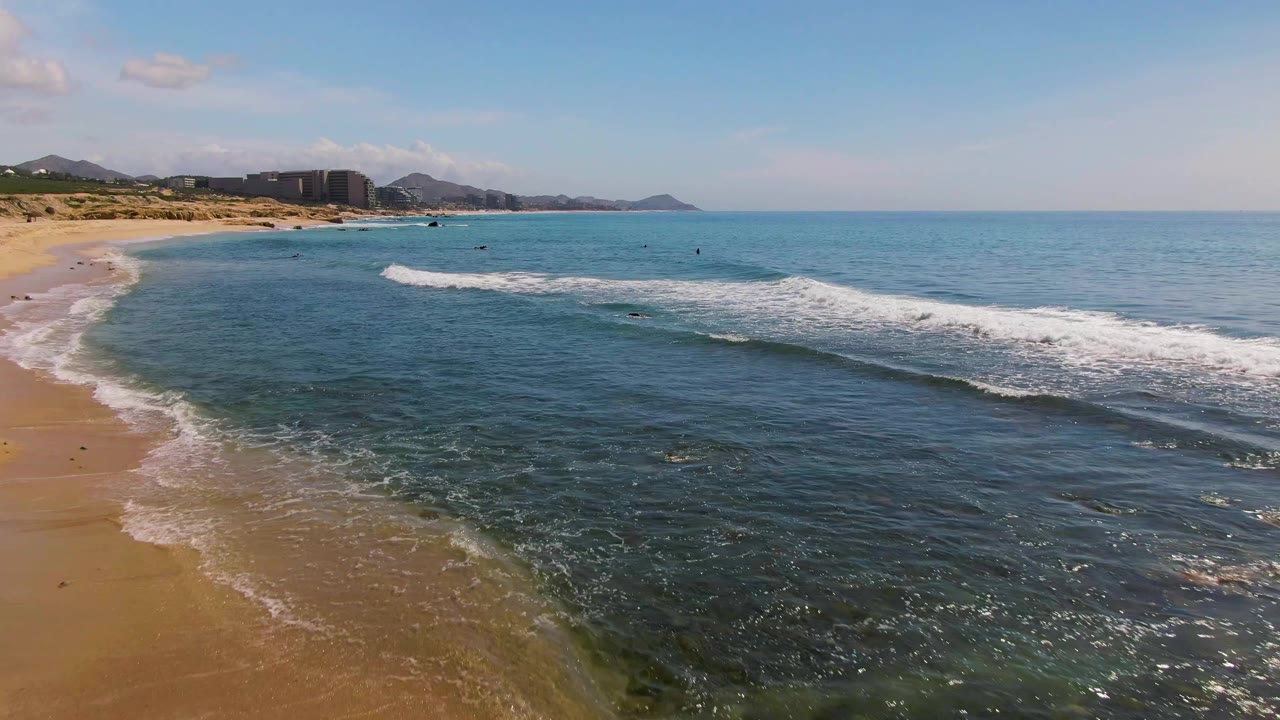 Aerial frame from El Tule Los Cabos with the shoreline closer to the left edge, white surf spreading across the middle water, distant buildings and hills, and no visible people.