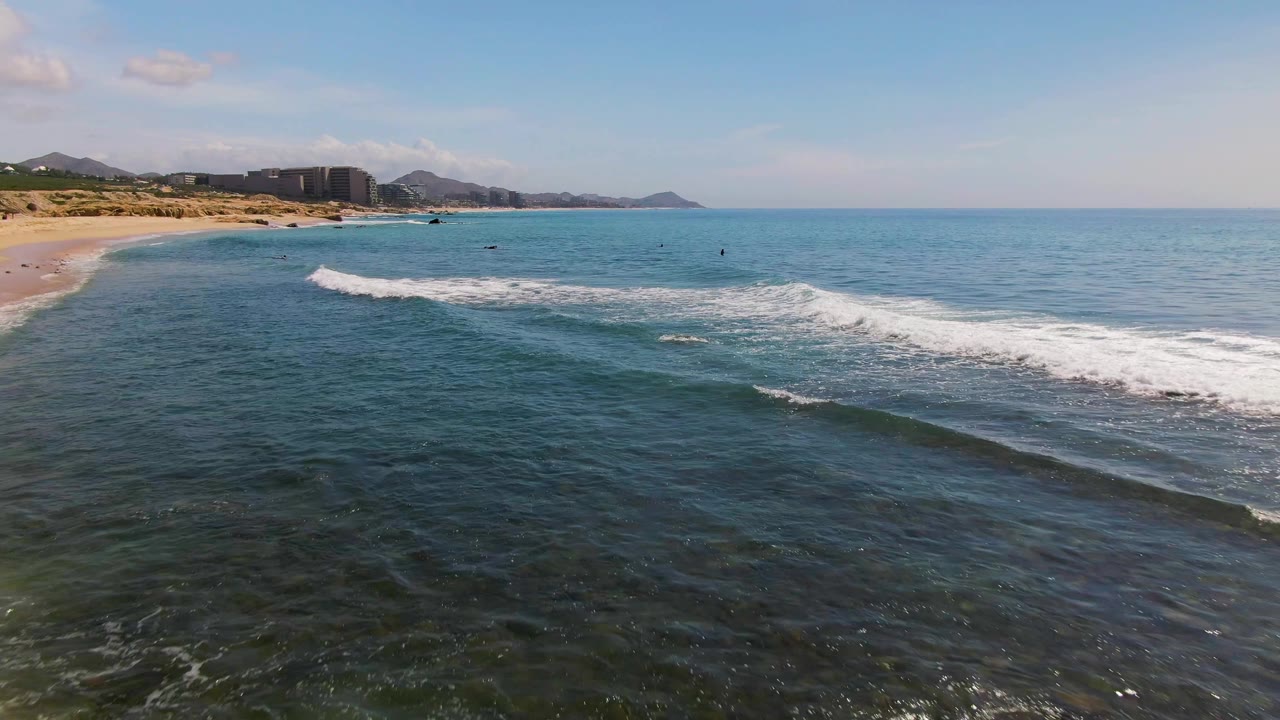 El Tule Los Cabos aerial view with darker foreground water, a broad white breaking wave near the right side, sandy beach and developed shoreline in the distance, and no visible people.