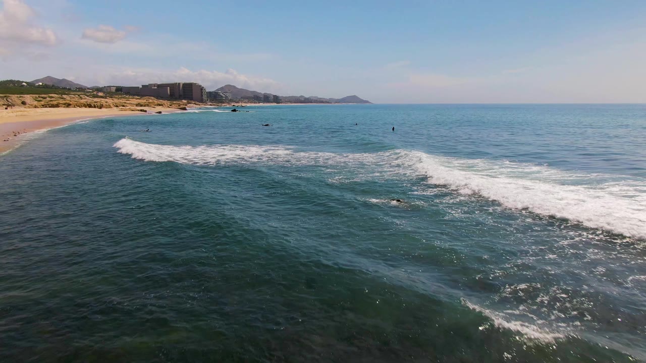 Wide aerial frame at El Tule Los Cabos with a curved sandy beach on the left, a foamy wave band across the center, calm open water to the right, distant hills, and no visible people.