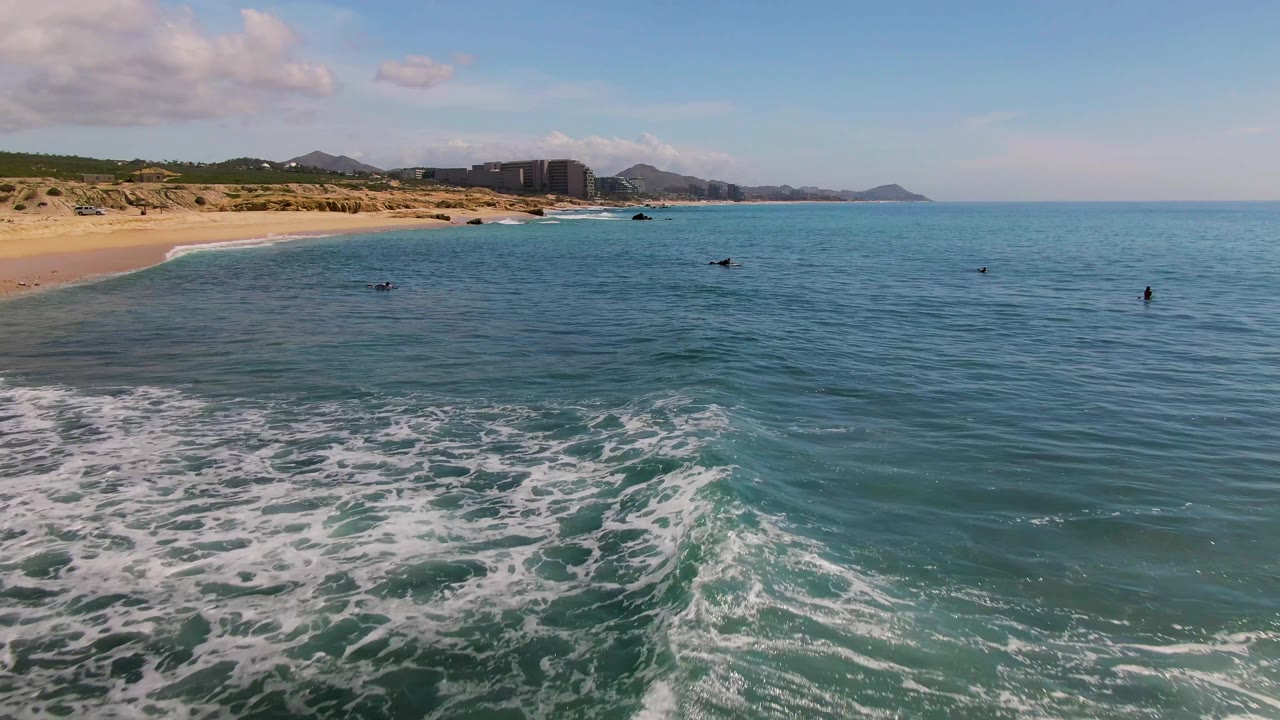 Aerial view at El Tule Los Cabos showing swimmers spaced across blue-green water, white foam in the foreground, and a sandy beach with distant hills and buildings.