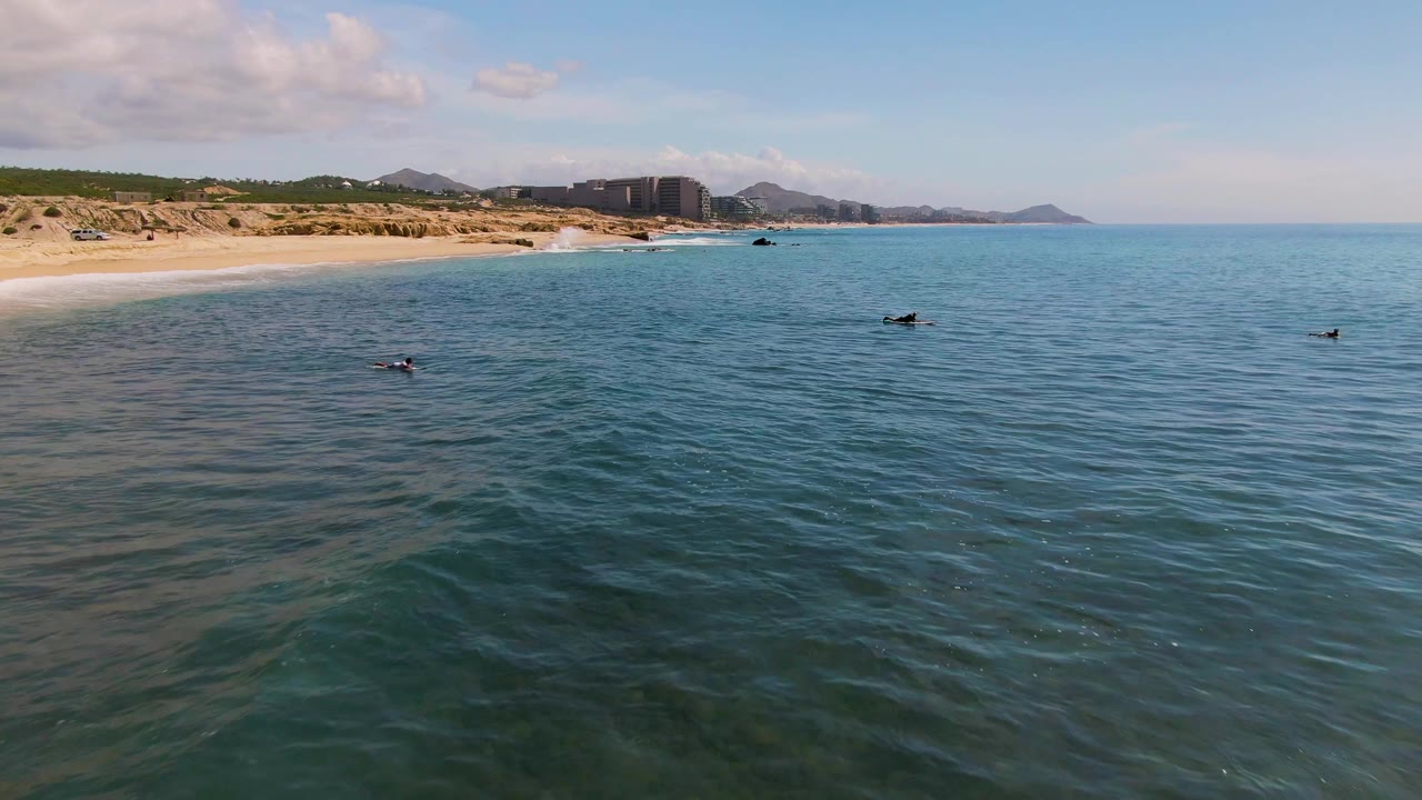 At 0:02, the El Tule Los Cabos shoreline sits high in frame while swimmers appear as small dark figures across the blue water under scattered clouds.