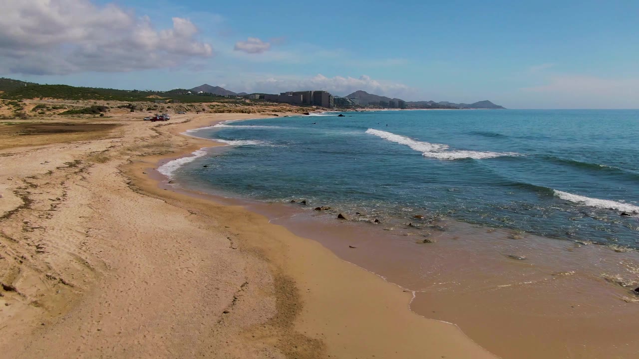Aerial view of El Tule Los Cabos beach with tan sand at left, blue ocean at right, small surf lines, distant buildings and mountains, and no visible people.
