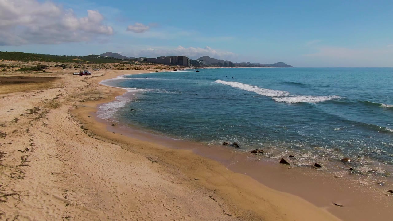 Elevated frame over El Tule Los Cabos showing the curving sandy shore, a white wave line offshore, scattered shoreline rocks, distant buildings, mountains, and no visible people.