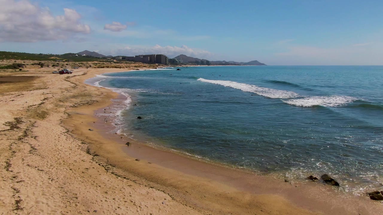 Wide aerial composition at El Tule Los Cabos with clear blue-green water in the foreground, sandy beach along the left, breaking surf near the middle, distant buildings, mountains, and no visible people.