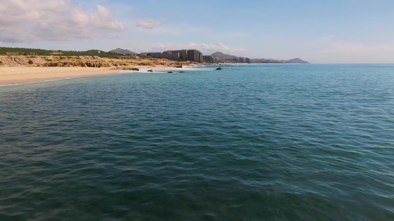 Opening frame of El Tule Los Cabos with blue-green ocean in the foreground, sandy beach to the left, distant buildings and hills on the horizon, and no visible people.