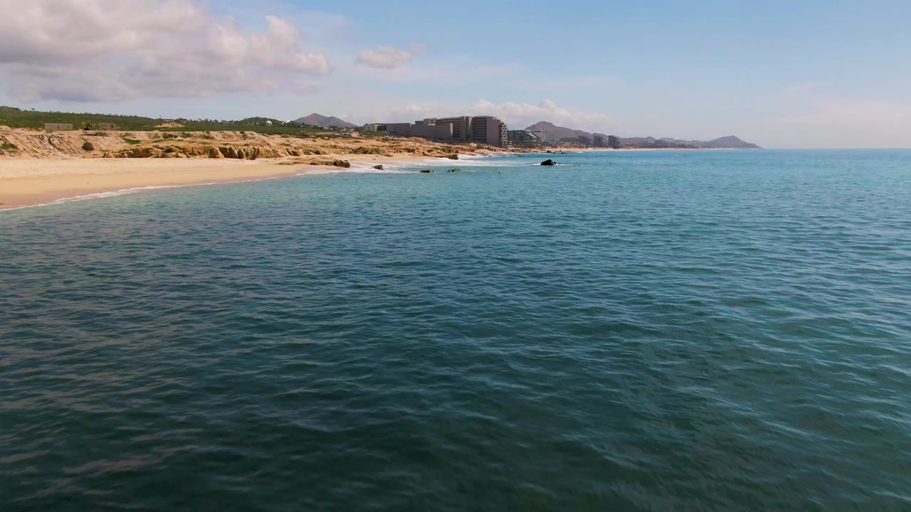 El Tule Los Cabos shoreline at 0:01 with rippled turquoise water across the foreground, pale sand and low cliffs at left, distant buildings, hills, and no visible people.