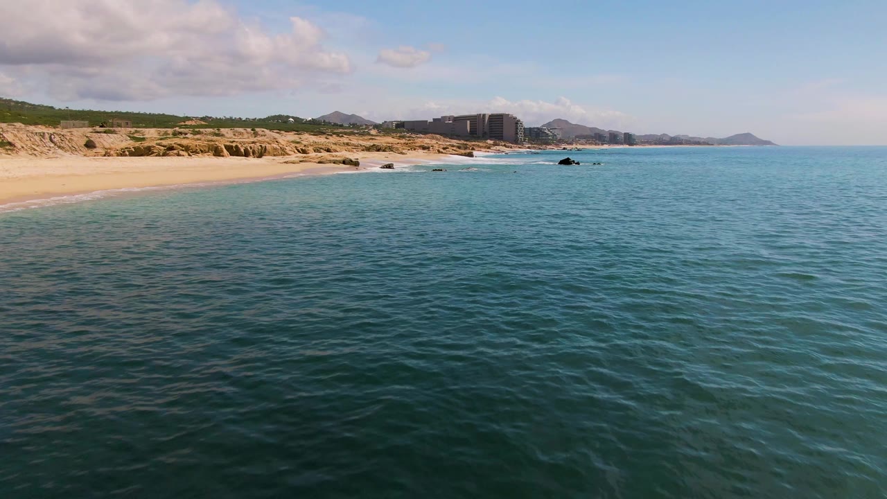 El Tule Los Cabos frame with darker rippled water in the foreground, a sandy beach and rocky edge to the left, distant beachfront buildings, low hills, and no visible people.