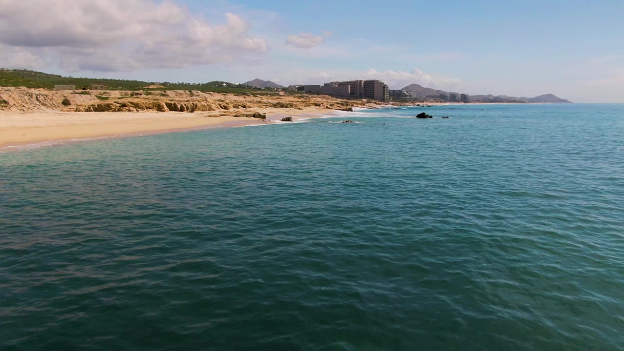 Ocean foreground with empty sand beach, low cliffs, rocks, buildings, and hills.