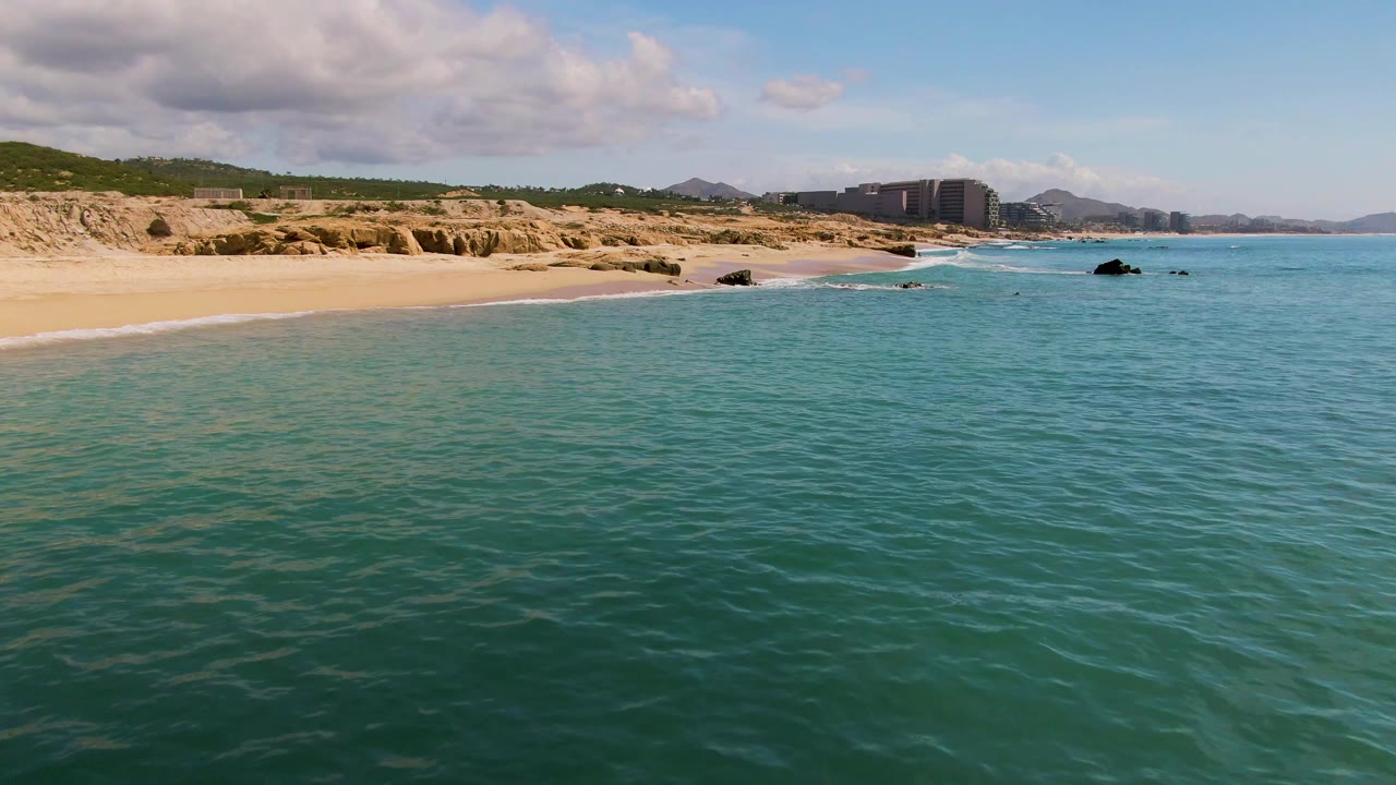 Aerial view of turquoise water meeting a sandy El Tule Los Cabos beach, with rocky cliffs, green hills, distant buildings, partly cloudy sky, and no visible people.