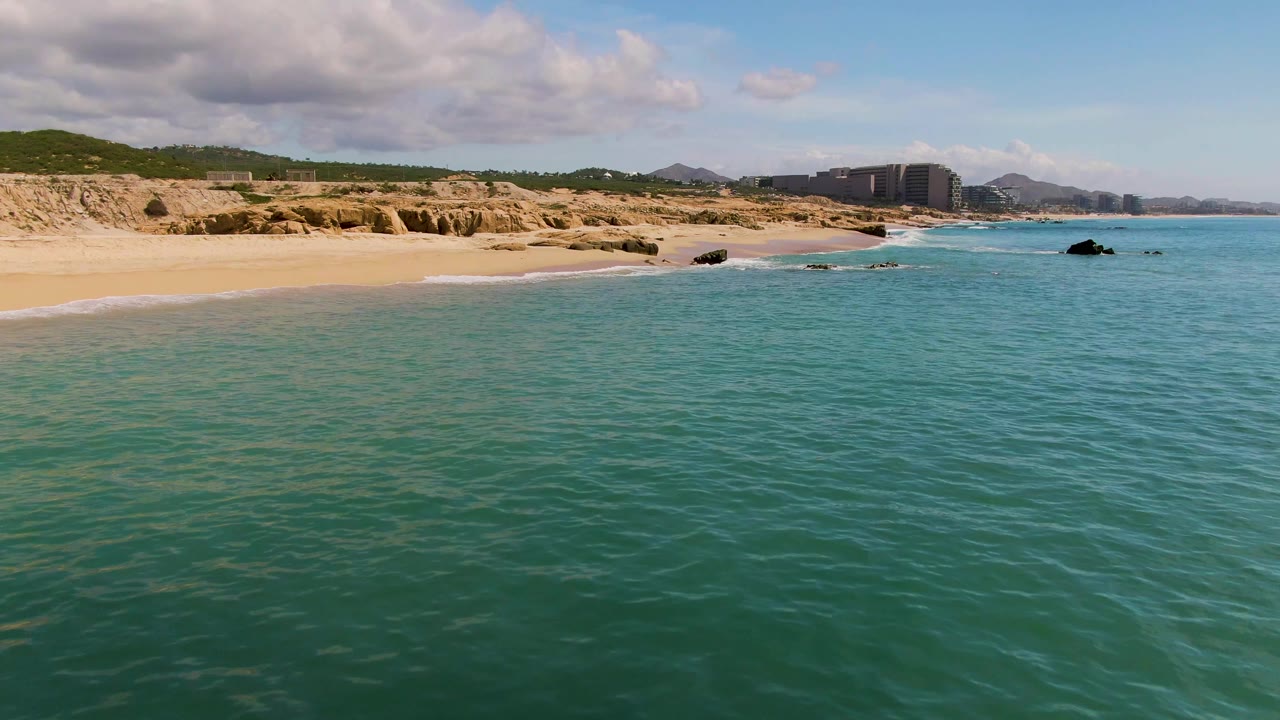 Clear turquoise water ripples toward the El Tule Los Cabos shoreline, with pale sand, low tan rock formations, green hills, distant buildings, and no visible people.