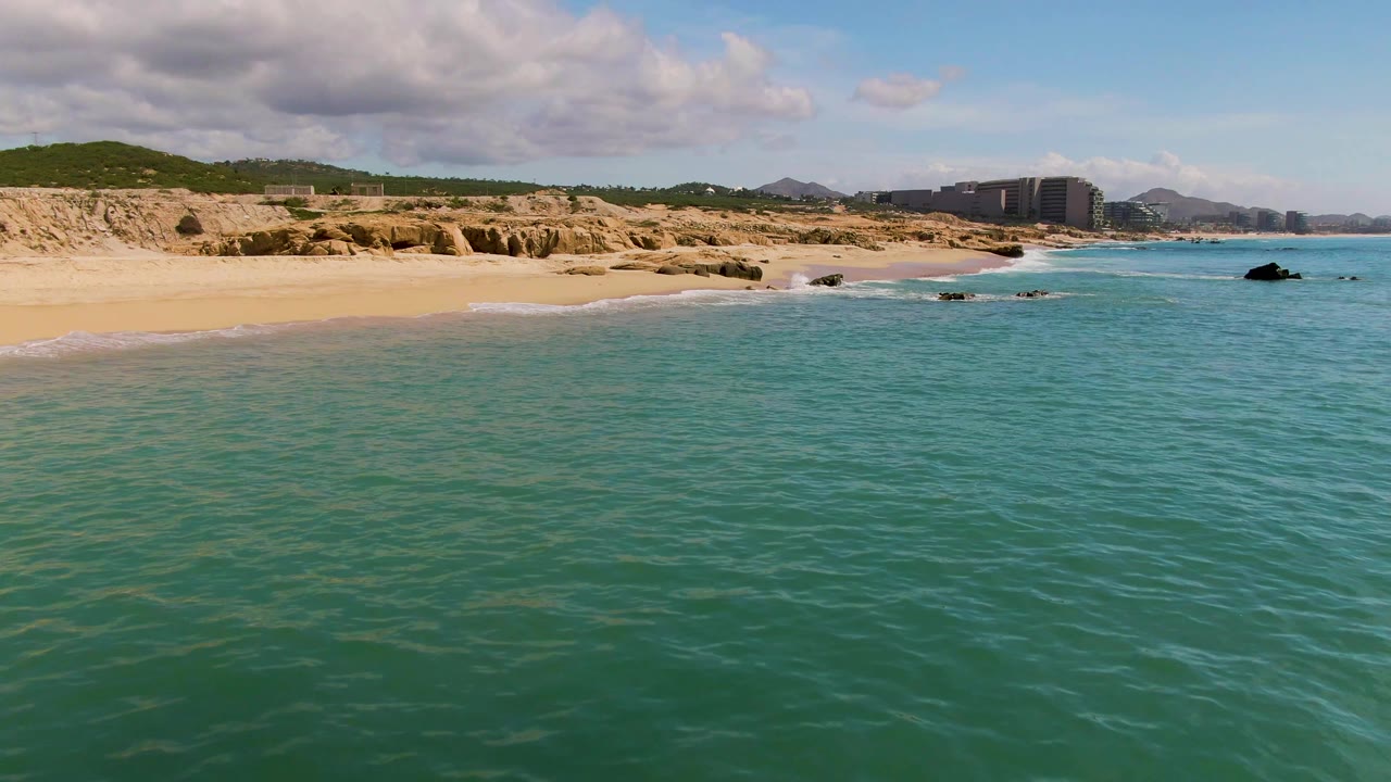 The El Tule Los Cabos beach appears slightly farther along the shore, with small waves touching sand, dark rocks offshore, low cliffs, distant buildings, and no visible people.