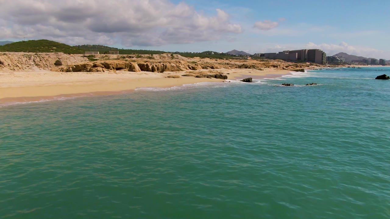 Wide coastal view with turquoise water, sandy beach, cliffs, hills, and clouds.
