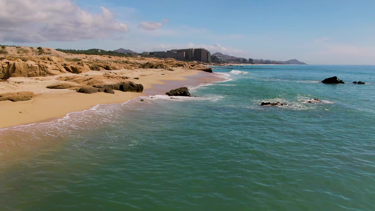 Elevated view of El Tule Los Cabos shoreline with turquoise water, tan sand, rocky outcrops, distant buildings, low hills, scattered clouds, and no visible people.