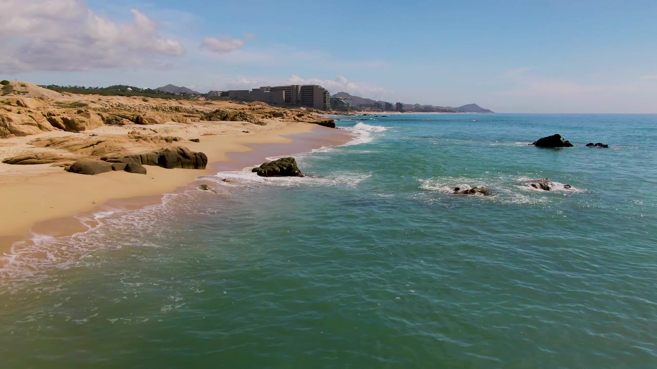 El Tule Los Cabos beach seen from above with water pushing farther along the sand, dark rocks in the surf, distant shoreline buildings, mountains, and no visible people.