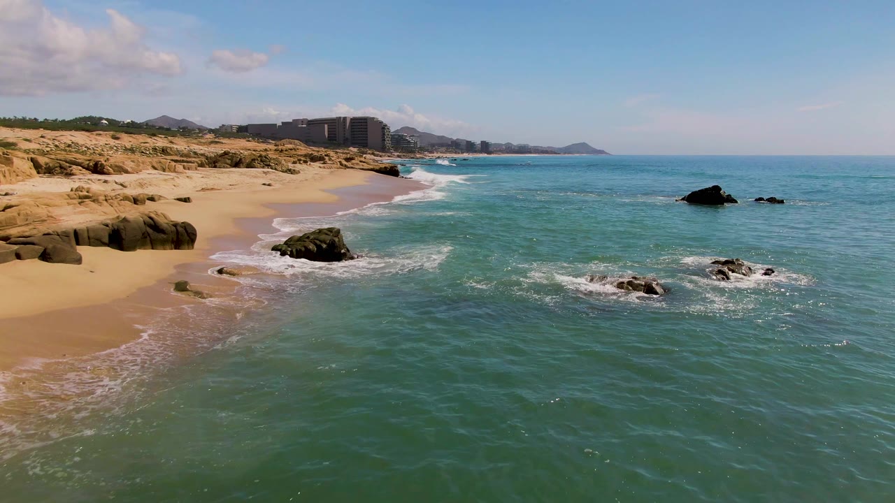 Wide elevated frame of El Tule Los Cabos with the curving sandy shore at left, turquoise water at right, clustered rocks breaking the surf, distant buildings, and no visible people.