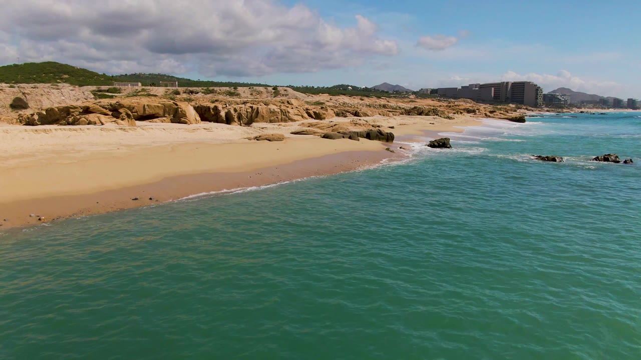 El Tule Los Cabos shoreline seen from above with shallow turquoise water, a narrow wave edge on the sand, tan coastal rocks, distant buildings, and no visible people.