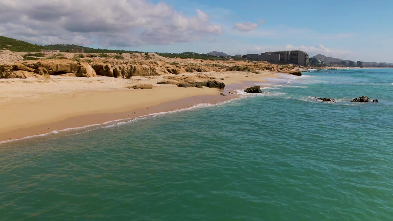 Wide aerial frame of El Tule Los Cabos showing textured turquoise water, pale sand, rocky shore ledges, distant coastal buildings, hills, and no visible people.