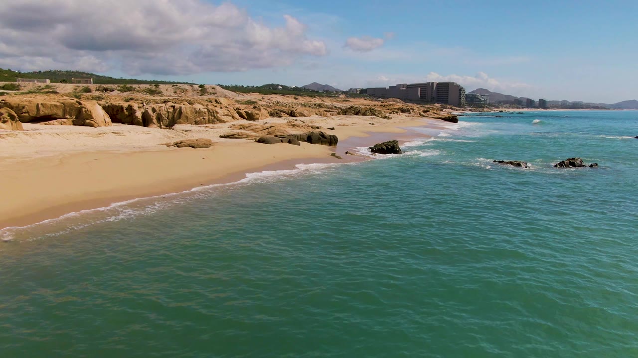 Aerial view over open turquoise water toward the sandy El Tule Los Cabos beach, with small waves near dark rocks, tan cliffs, distant buildings, and no visible people.