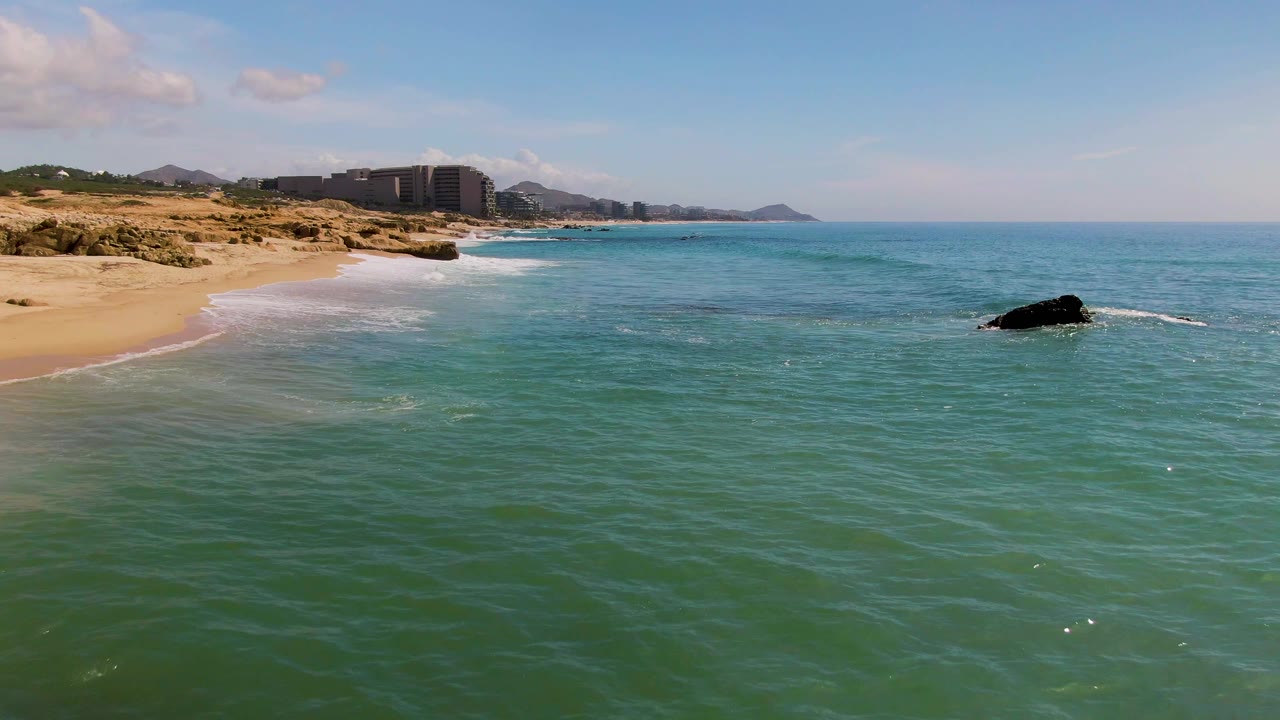 Green-blue water spreads across the foreground at El Tule Los Cabos, with white surf along the sandy beach, the offshore rock farther right, and no visible people.