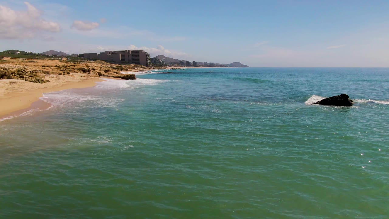 The El Tule Los Cabos shoreline sits along the left edge while the offshore rock shifts closer to the right side of frame, with low surf and no visible people.