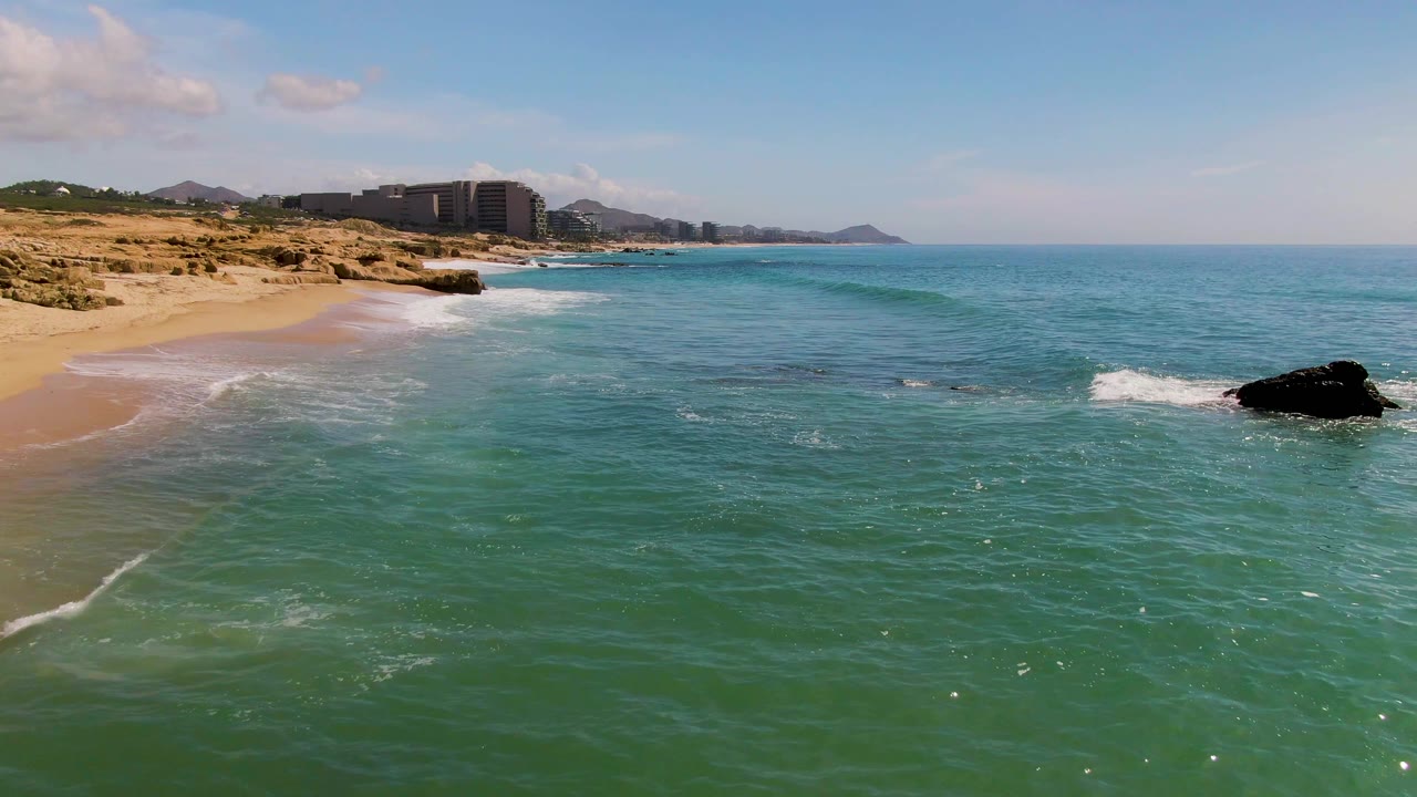 At El Tule Los Cabos, a thin line of foam reaches the sand while green-blue water fills the frame and the dark offshore rock sits near the right edge, with no visible people.