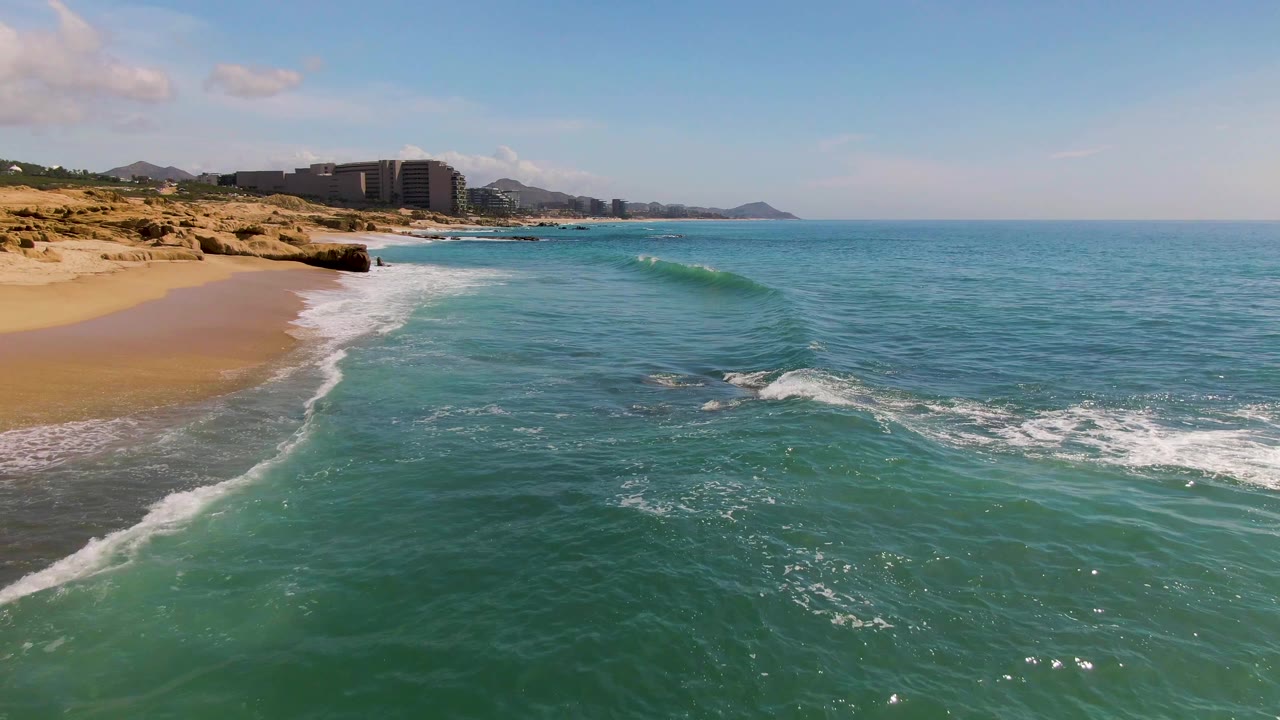 Opening frame over El Tule Los Cabos shows turquoise water rolling toward tan sand, rocky shoreline at left, distant buildings and hills, blue sky, and no visible people.