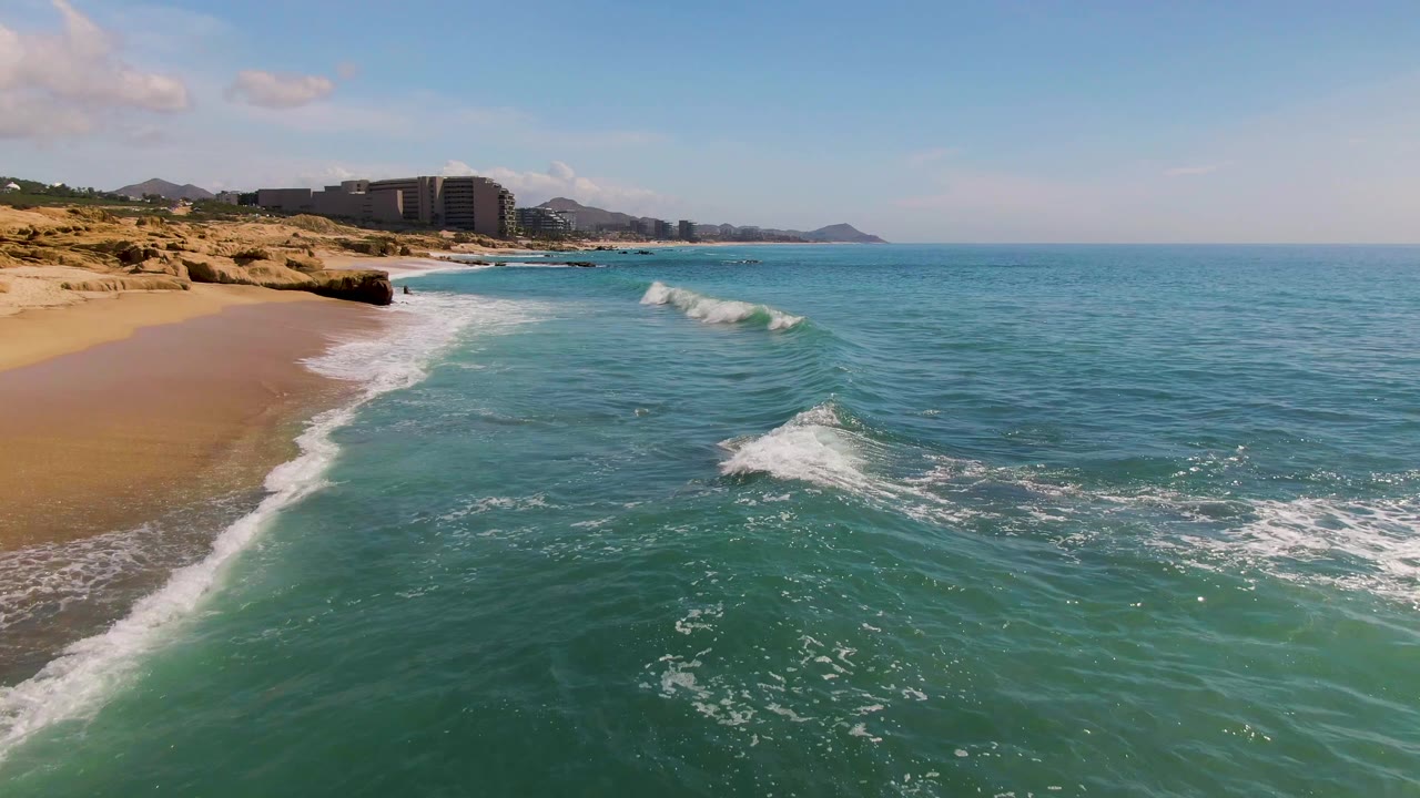At 0:01 in El Tule Los Cabos, a low wave begins to crest offshore while a white foam line traces the sand, with distant buildings under clear blue sky and no visible people.