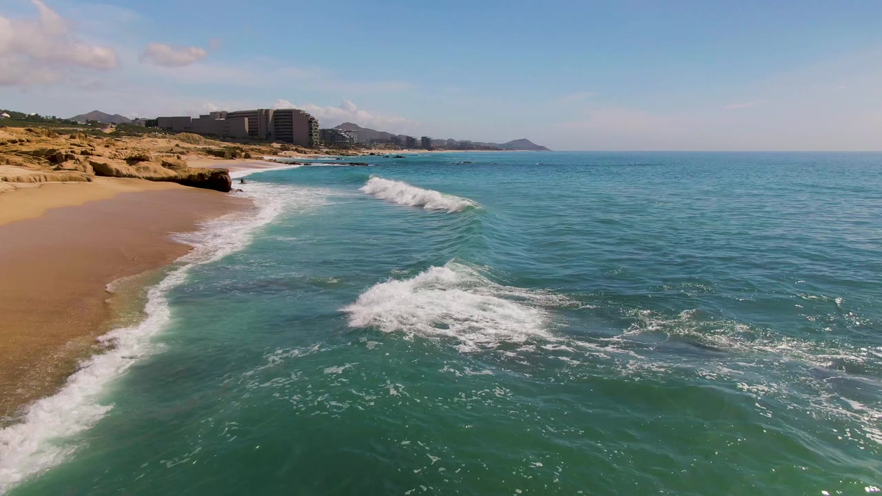 At 0:02 in El Tule Los Cabos, the wave breaks into a brighter patch of white foam in the midground, with open turquoise water to the right, beach at left, and no visible people.