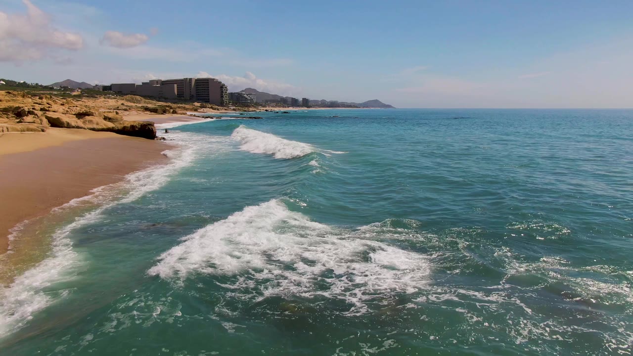 Foam spreads across turquoise water beside a sandy shoreline and rock outcrop.
