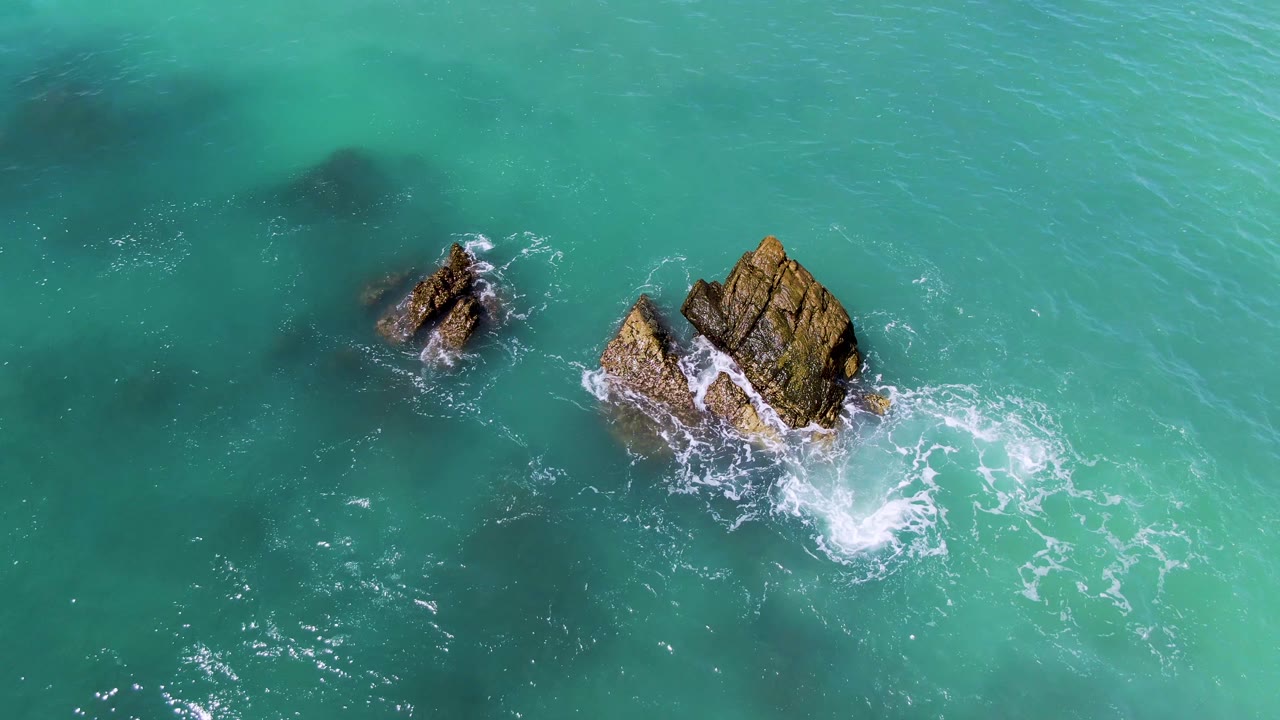 Overhead view at El Tule Los Cabos showing two rocky outcrops in turquoise water, white foam curling around the larger rock, and no visible people.