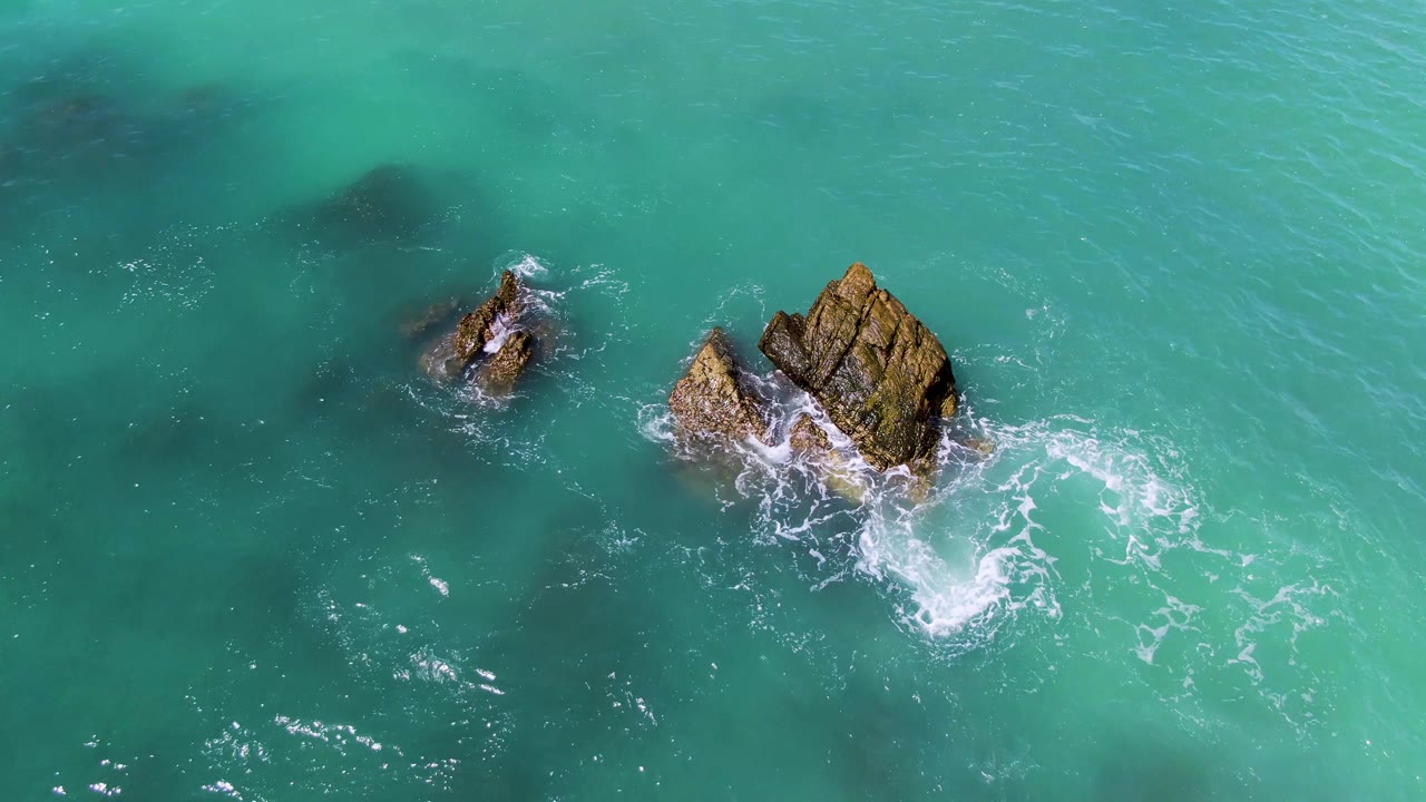 Aerial frame at El Tule Los Cabos with the larger rock near the right side, a smaller rock to the left, spreading white foam, and no visible people.