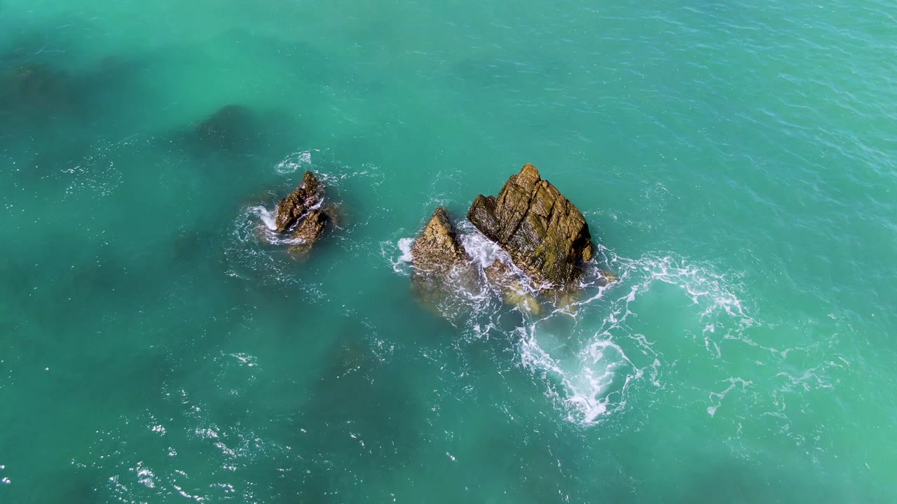 Aerial turquoise sea with rocks and foam shifted below the outcrops.