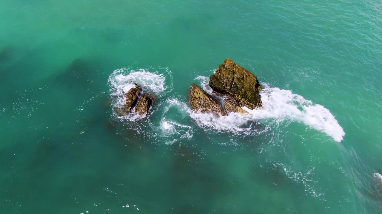 At 0:01, waves push white foam away from the larger rock in El Tule Los Cabos, with darker underwater features visible and no visible people.