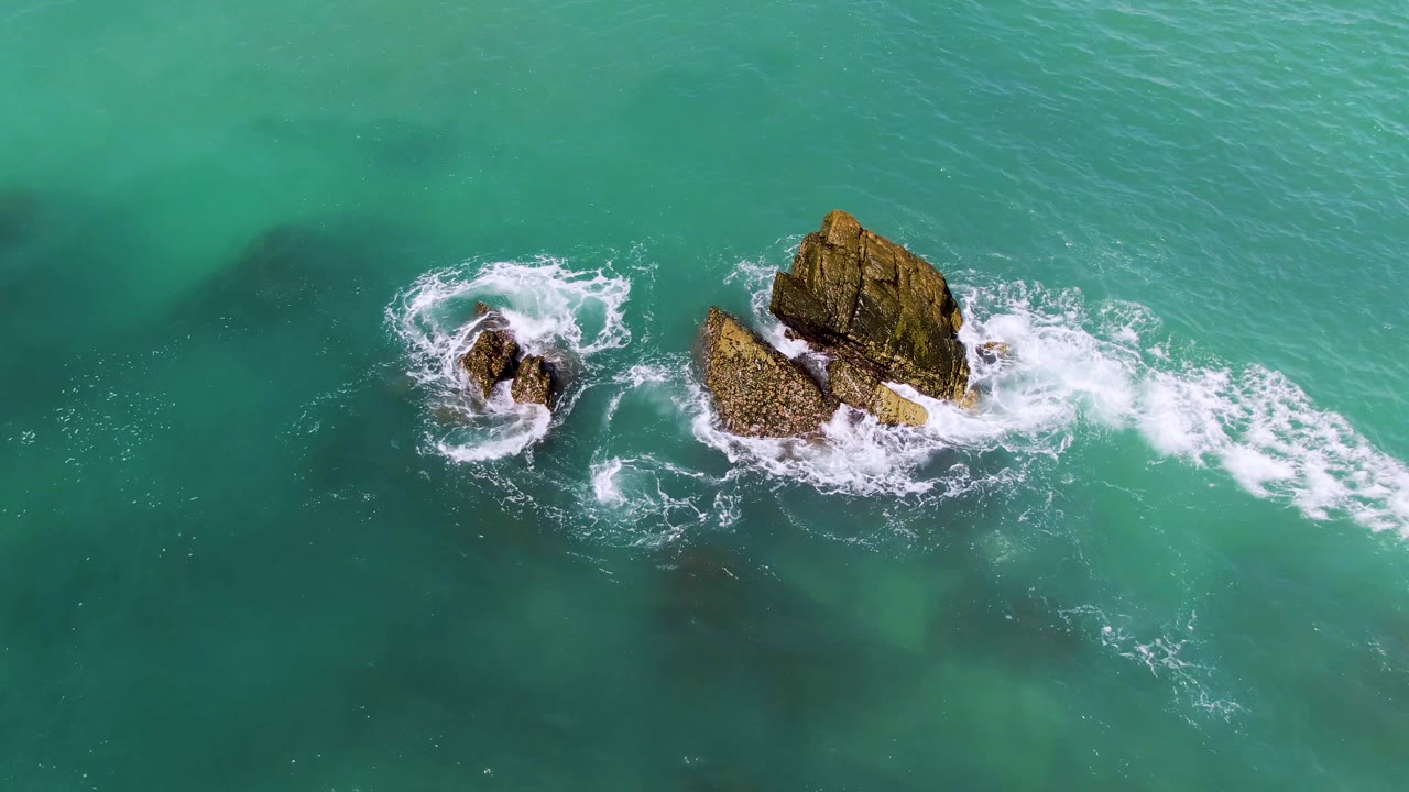 At 0:02, the surf wraps around the smaller rock and breaks against the larger outcrop in El Tule Los Cabos, with open turquoise water around them and no visible people.
