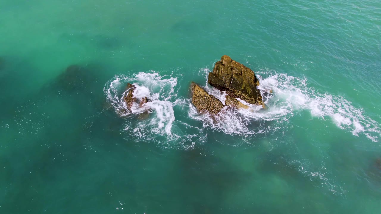 At 0:03, white foam curls in a broad arc around the left rock while the larger outcrop sits in clear turquoise water at El Tule Los Cabos, with no visible people.