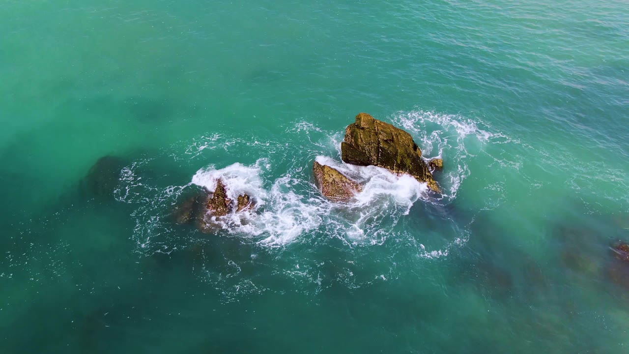 Overhead view at El Tule Los Cabos showing turquoise water and white foam circling two rocky outcrops, with no visible people.