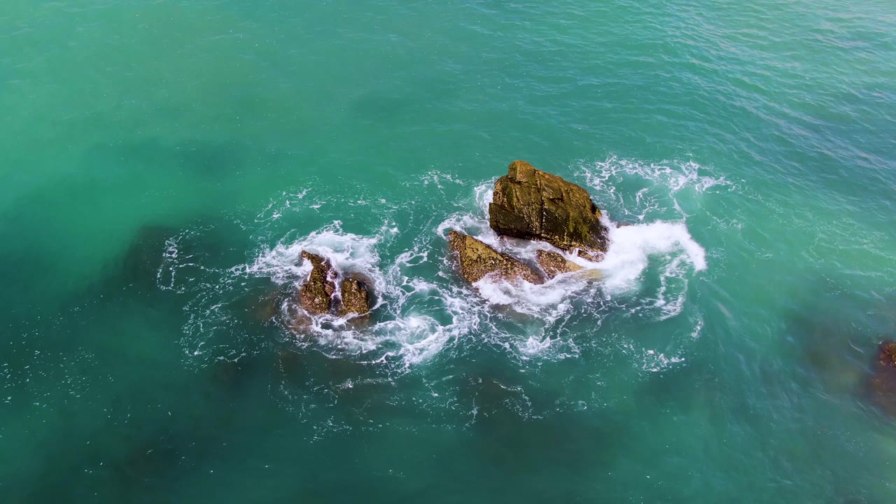At El Tule Los Cabos, white surf spreads wider around the central rock pair in green-blue water, with darker seabed patches visible and no people in frame.