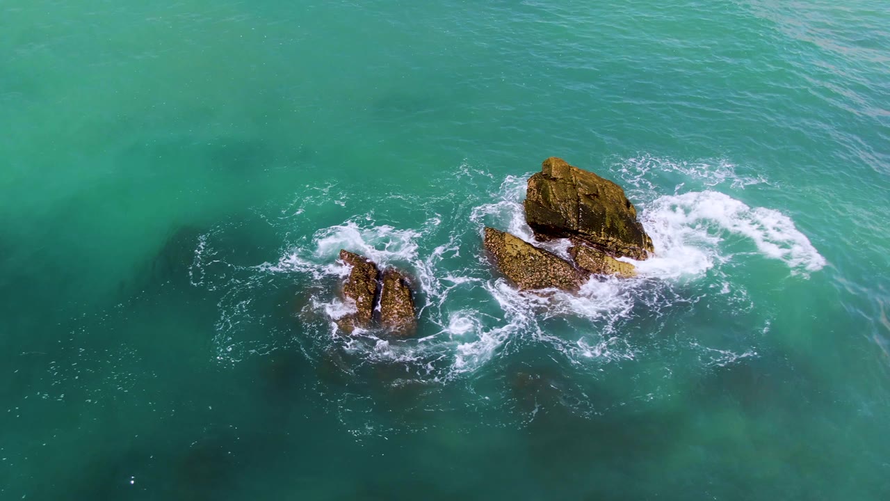 Overhead drone view at El Tule Los Cabos with waves breaking on two isolated rocks and clear turquoise water filling the frame, with no visible people.