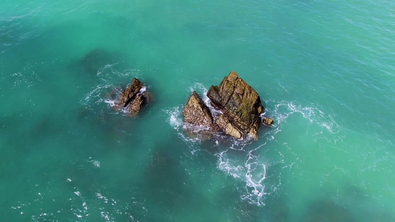 Overhead view of two rocky outcrops in turquoise water at El Tule Los Cabos, with light white foam around the larger rock and no visible people.