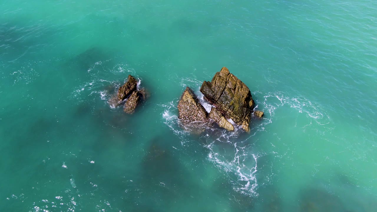 Two rock formations sit near the center of turquoise ocean water at El Tule Los Cabos, with small foam lines starting around the larger outcrop and no visible people.