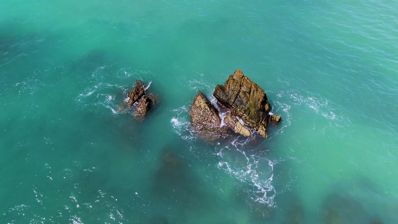 At El Tule Los Cabos, turquoise water surrounds the two rocky outcrops as white foam trails from the lower side of the larger rock, with no visible people.