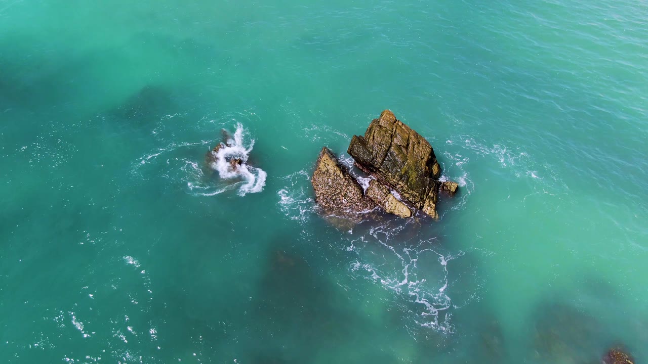 Wave splash breaks against the smaller rock beside a larger outcrop.