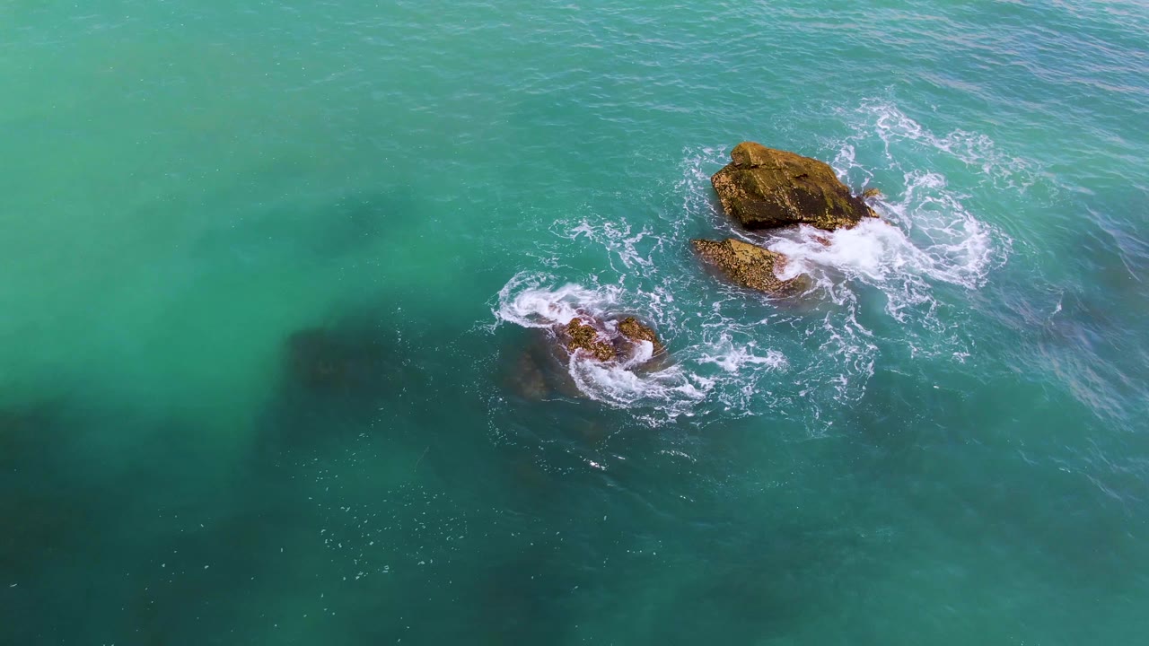 Aerial frame from El Tule Los Cabos with the larger rock near the upper right, a smaller rock below left, foam spreading outward, and no visible people.