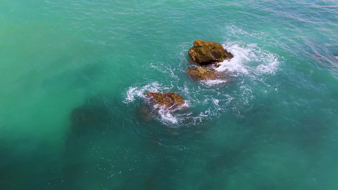 Aerial frame at El Tule Los Cabos where the smaller rock sits lower left, the larger rock sits upper center, white foam gathers around both, and no visible people appear.