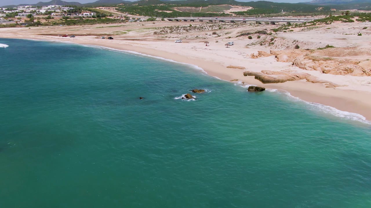 Aerial frame of El Tule Los Cabos showing turquoise ocean in the foreground, a pale sandy beach with rocky outcrops near the surf, distant vehicles by a road, and no visible people.