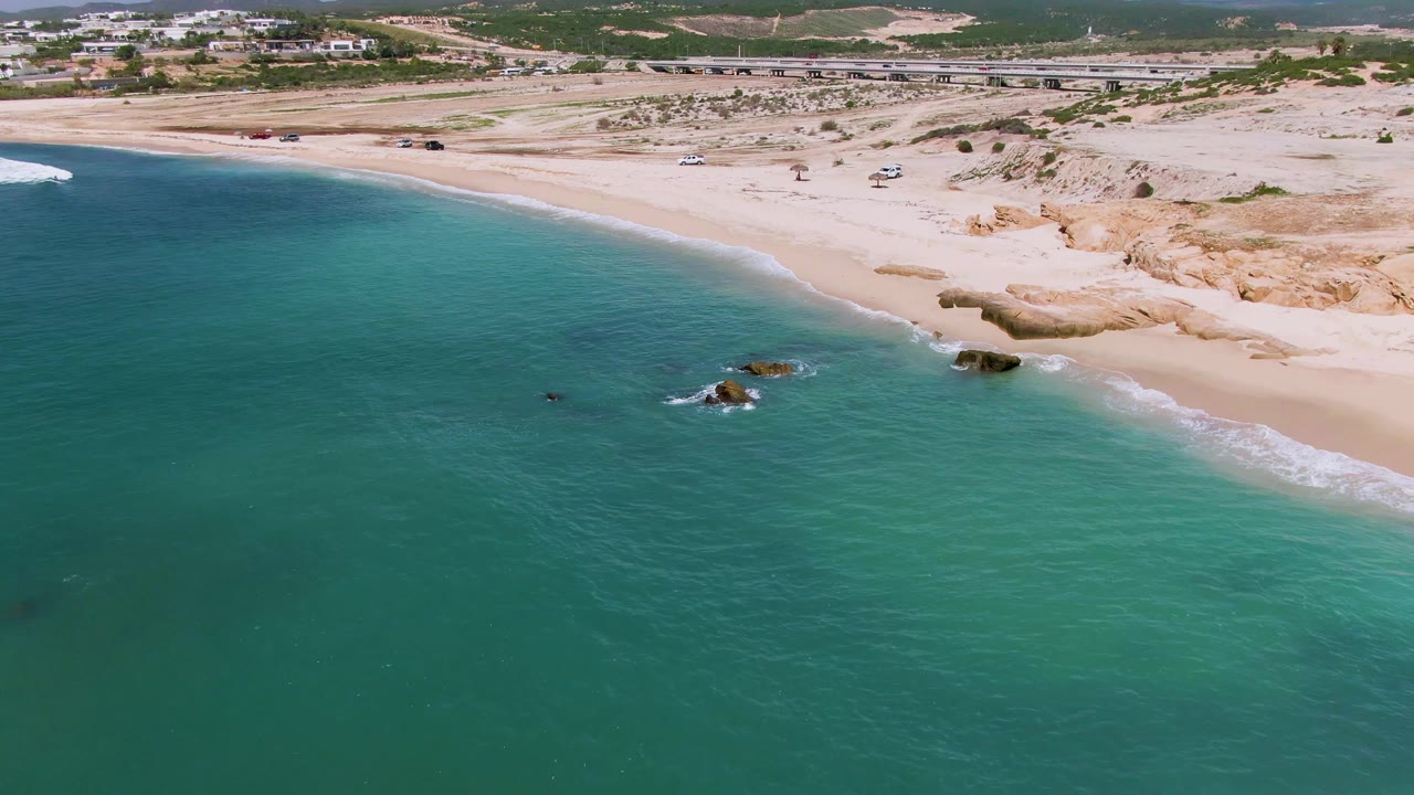 Aerial view over El Tule Los Cabos with the shoreline running diagonally, small rocks at the waterline, clear green-blue water, beach sand, distant road traffic, and no visible people.