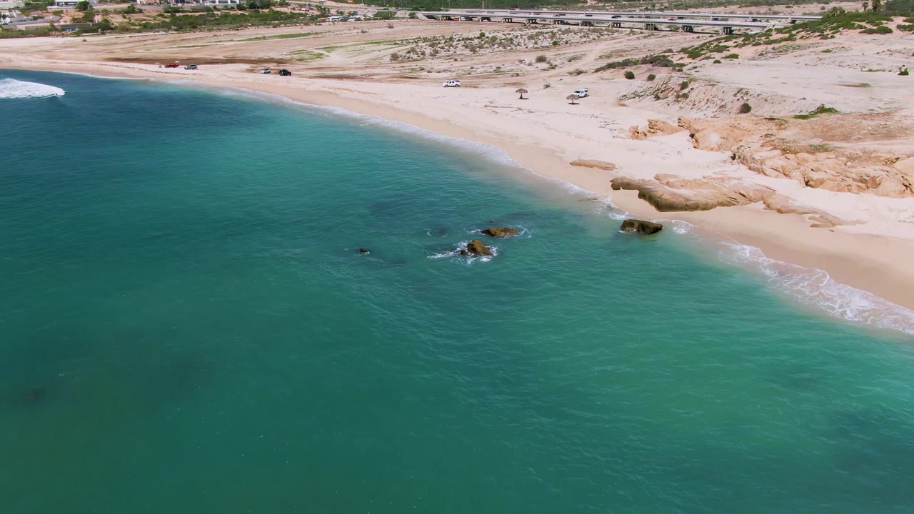 Wide aerial frame of El Tule Los Cabos with open turquoise water filling most of the view, rocky outcrops near the beach edge, pale sand, a background road with vehicles, and no visible people.