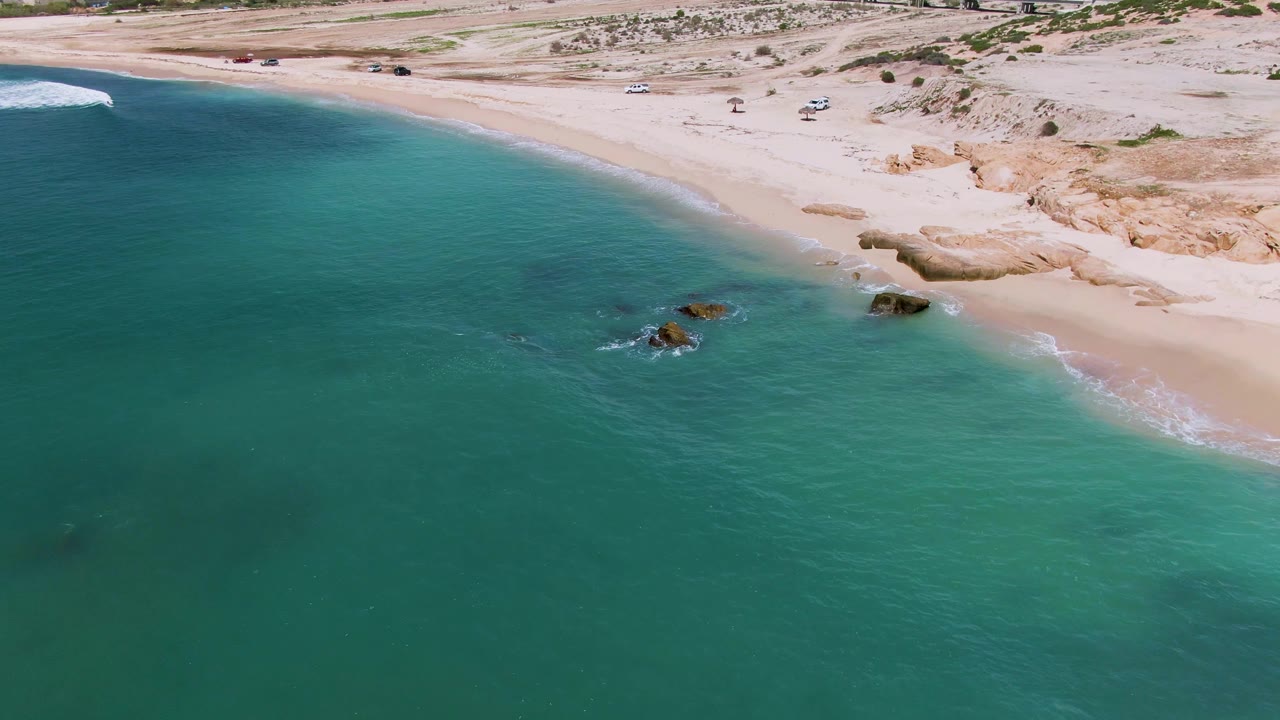 Aerial coastline with turquoise water, surf, rocks, beach sand, and sparse vehicles inland.